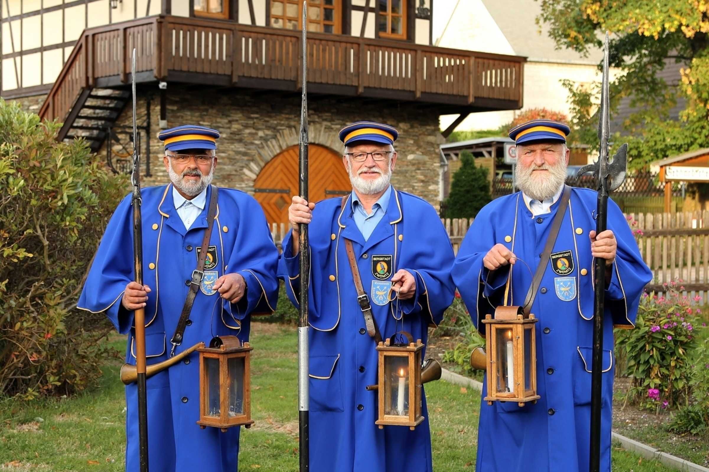 Drei Personen in blauen Uniformen halten Laternen und ein Gewehr, draußen vor einem Holzbauten.
