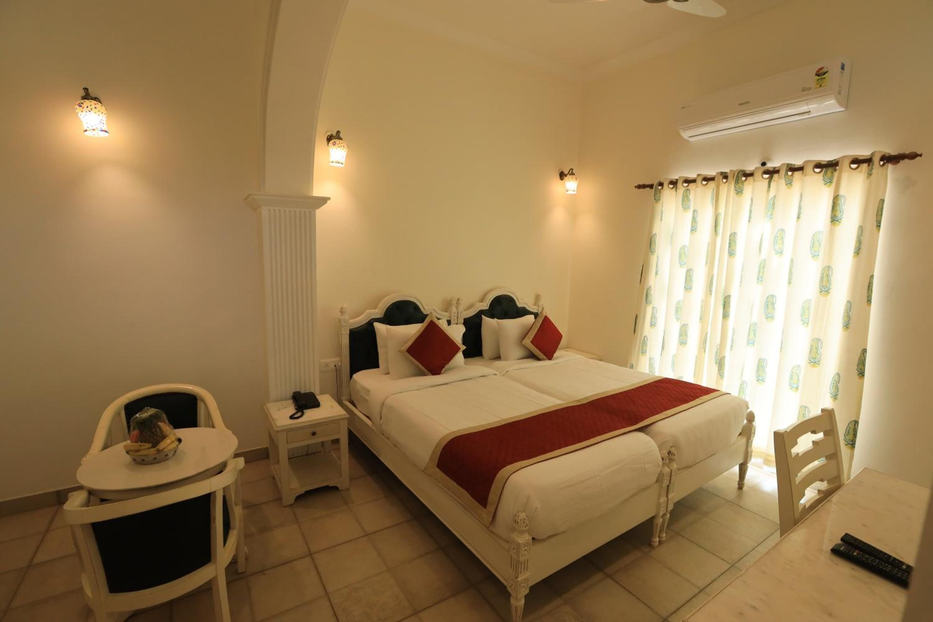 A hotel room with a bed, bedside tables, a chair, a small table, and an air conditioning unit.