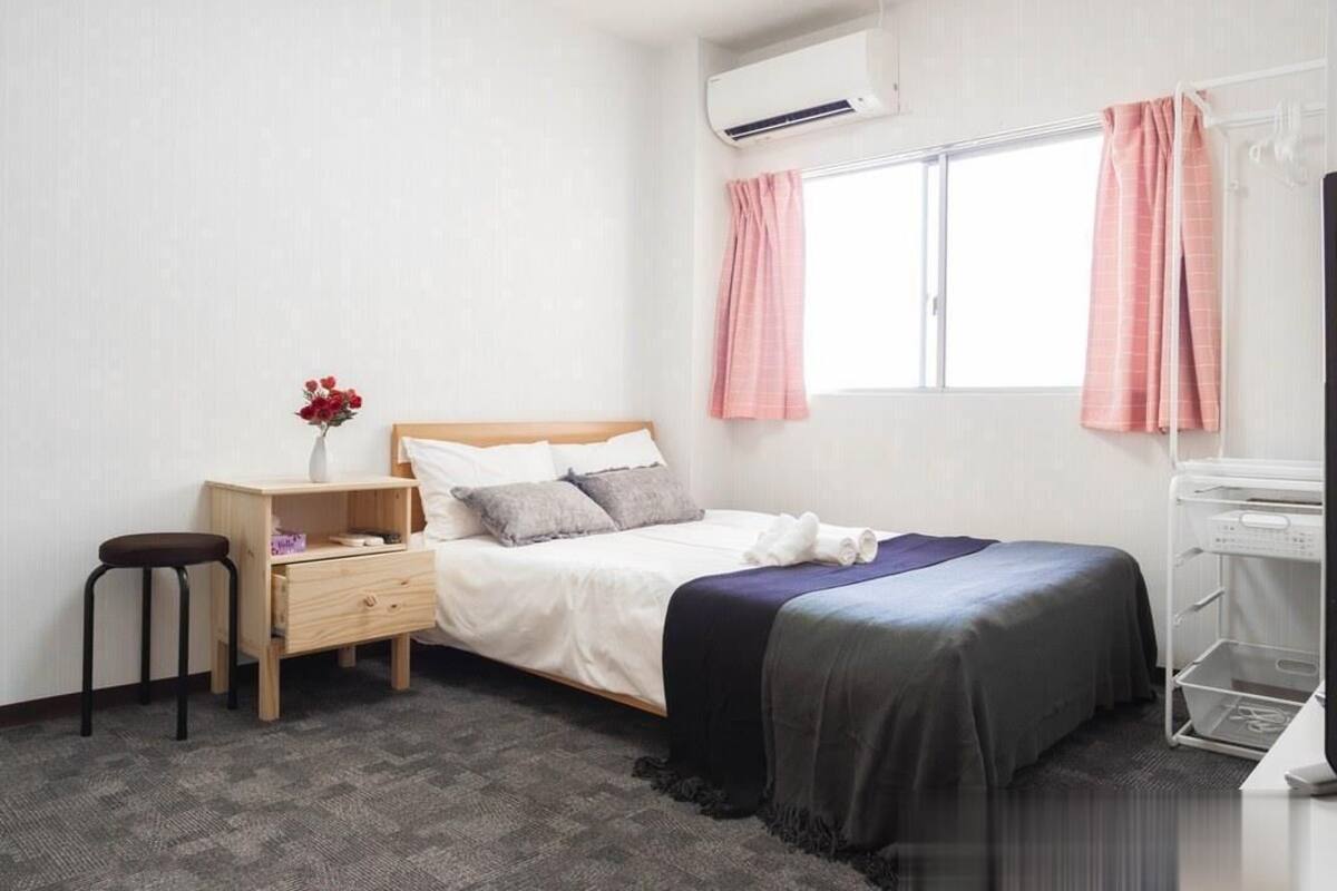 A bedroom with a bed, a nightstand, a chair, a window with curtains, an air conditioning unit, and a laundry cart.