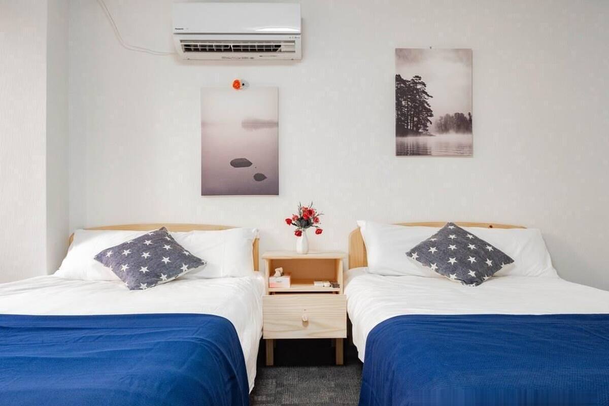 A hotel room with two beds, a nightstand, an air conditioner, and two wall-mounted artworks.