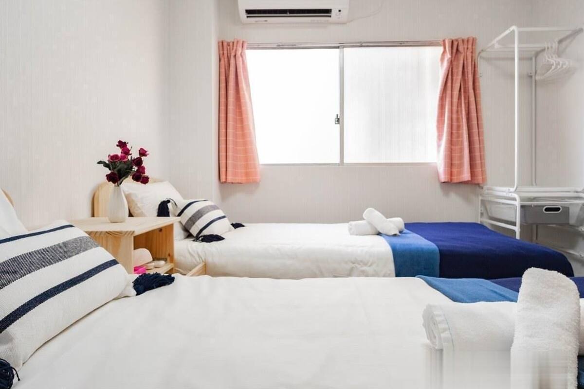 A hotel room with two beds, a small table with a vase of flowers, a window with curtains, and a white air conditioning unit.