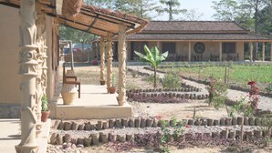 A traditional building with a covered porch, a garden with raised beds, and a large tree in the center. - Tharu Community Lodge (Kawasoti)