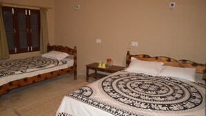 A room with two wooden beds, each with a patterned bedspread and white pillows. There is a small wooden nightstand between the beds with a lamp and a small table with a candle and a jar. - Tharu Community Lodge (Kawasoti)