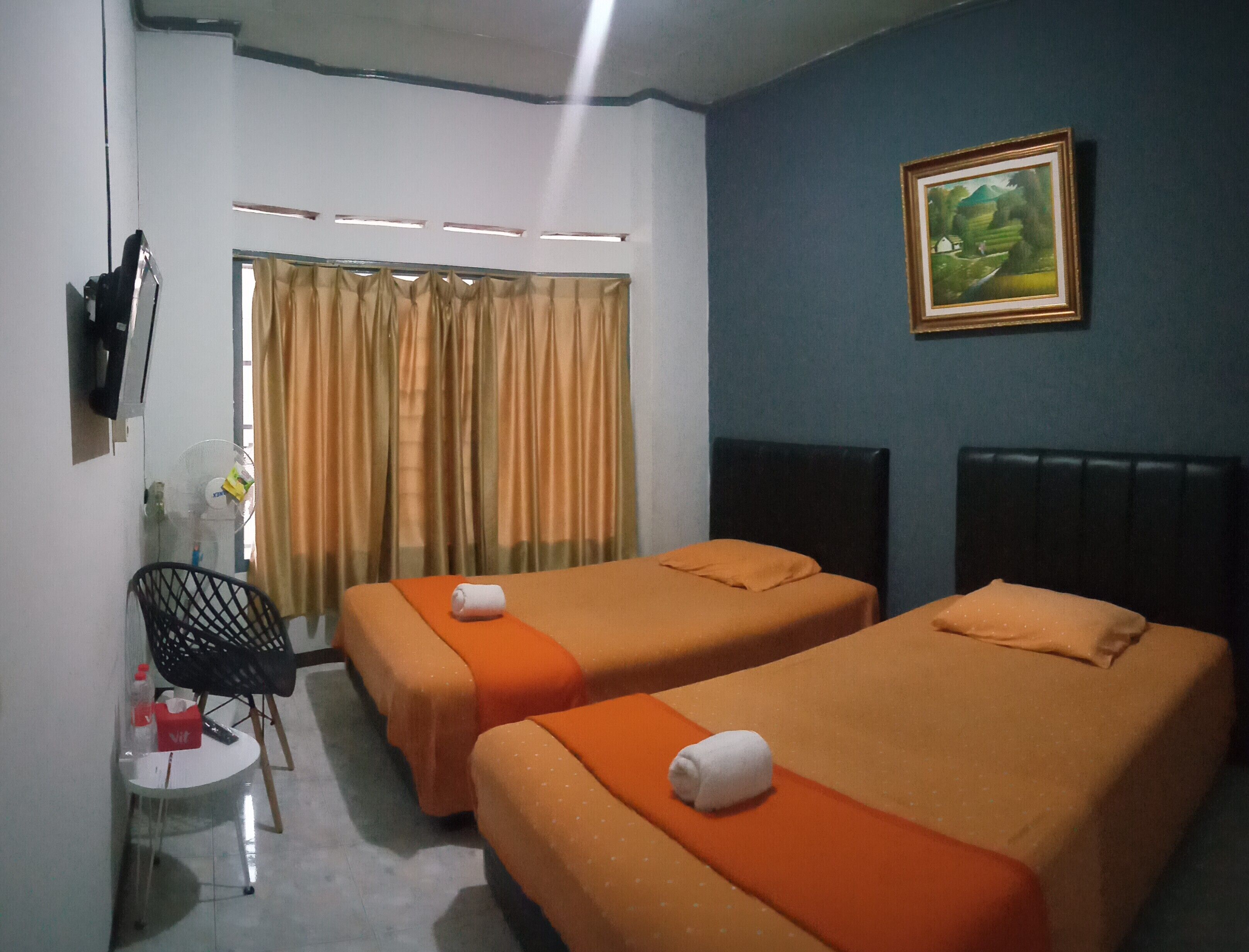A hotel room with two single beds, a small table with a chair, a TV mounted on the wall, and a window with curtains.