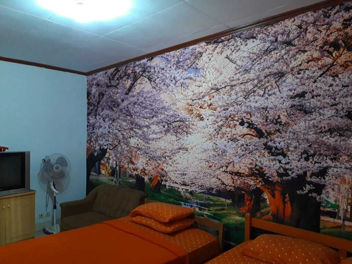 Una habitación con un gran mural en la pared de flores de cerezo, una cama con ropa de cama naranja, un televisor sobre un mueble de madera y un ventilador de pie.