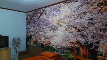 Una habitación con un gran mural en la pared de flores de cerezo, una cama con ropa de cama naranja, un televisor sobre un mueble de madera y un ventilador de pie.