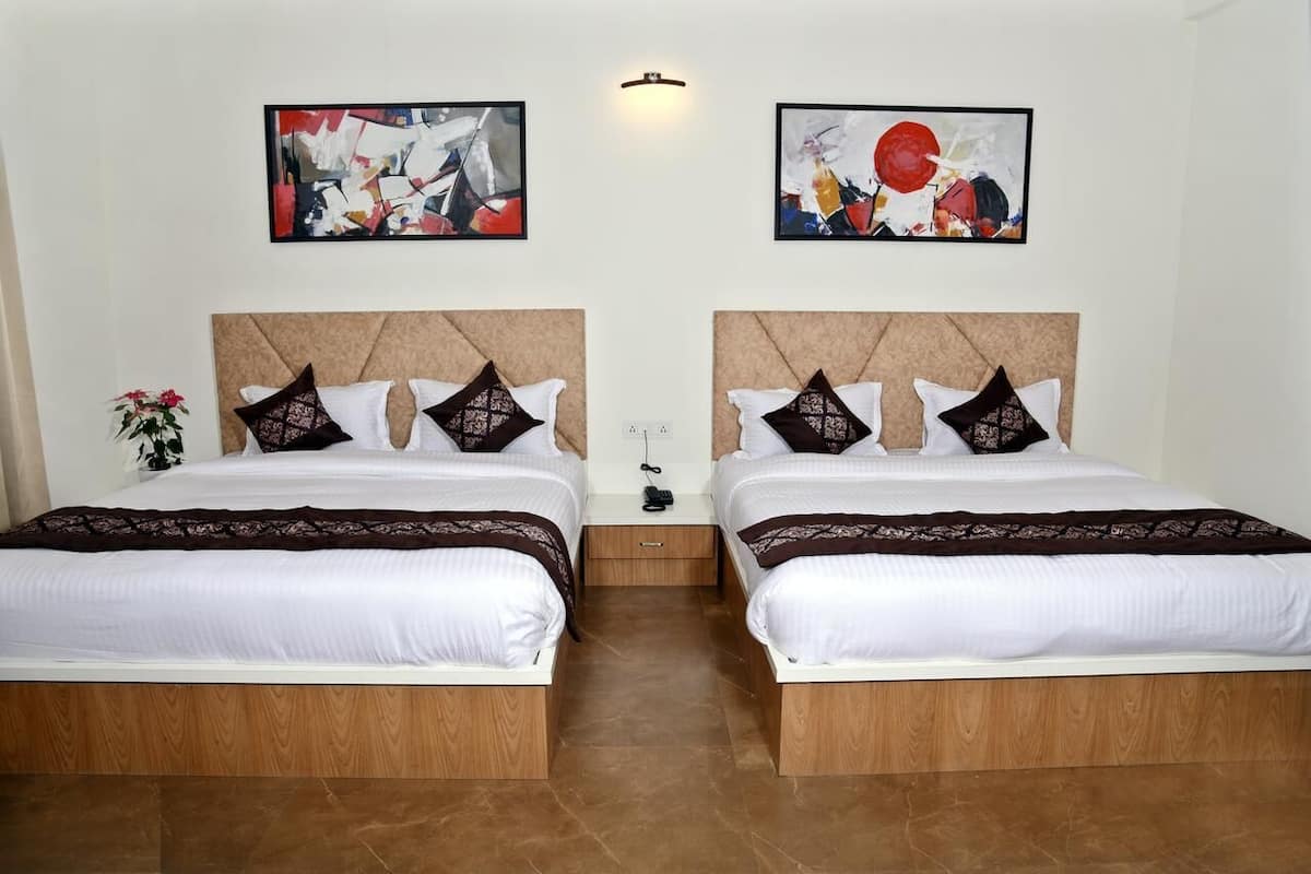 A hotel room with two beds, each with a headboard, white bedding, and decorative pillows. There are two abstract paintings on the walls, a small plant on the left side, and a wall-mounted light fixture above the beds.