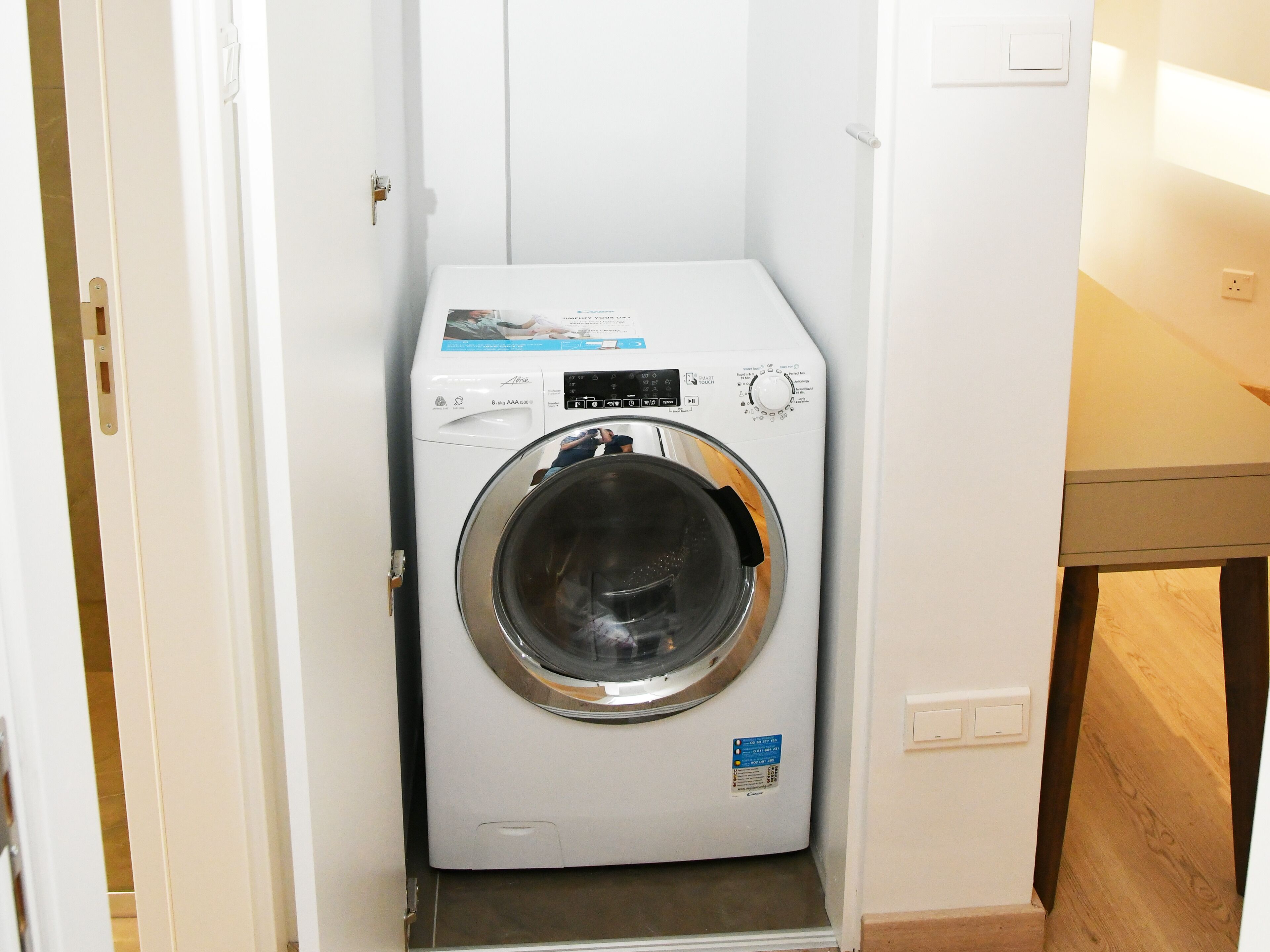 A compact, white front-loading washing machine in a small laundry room with a wooden floor and white walls.