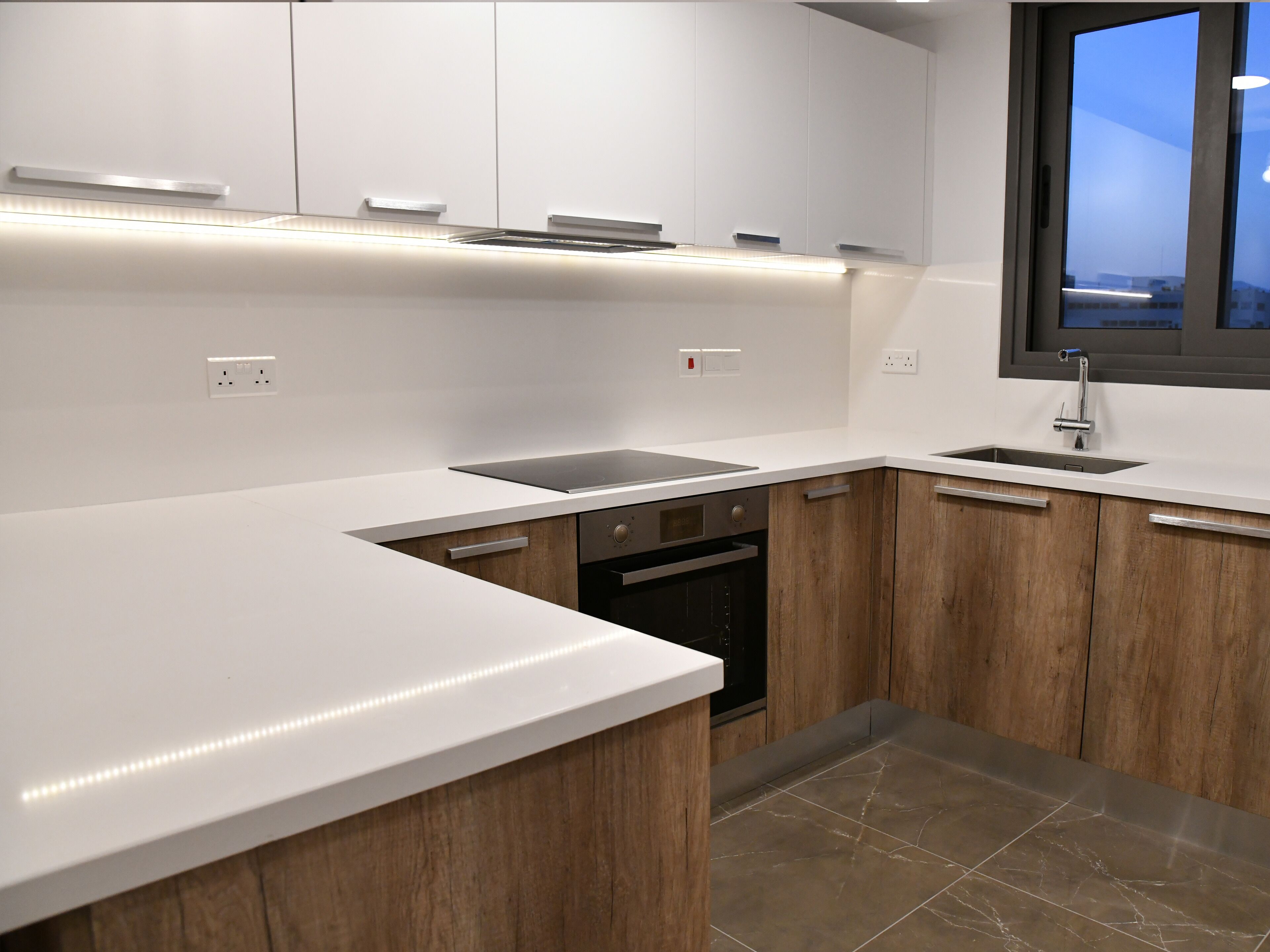 Una cocina moderna con encimeras blancas, armarios de madera, horno empotrado y ventana con vistas.