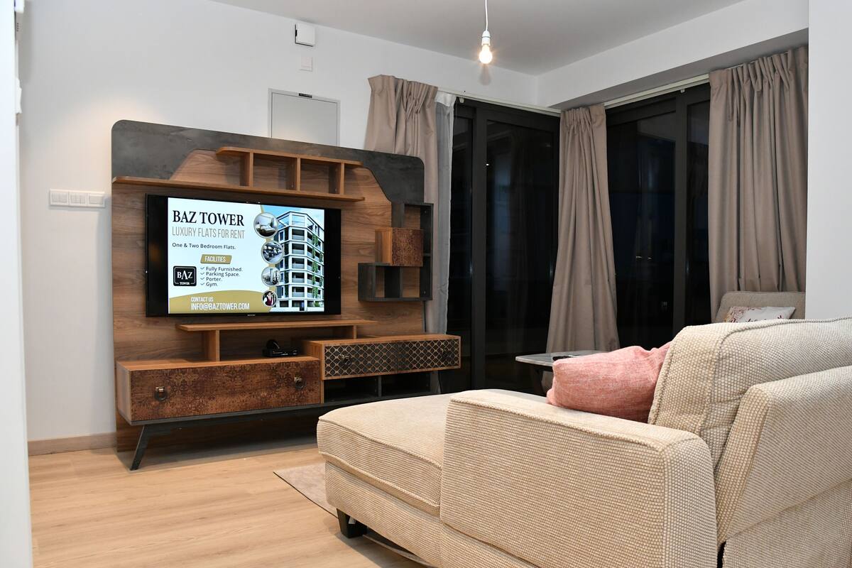 A modern living room with a flat-screen TV mounted on a wooden wall, a beige sofa, and a small table with a lamp.
