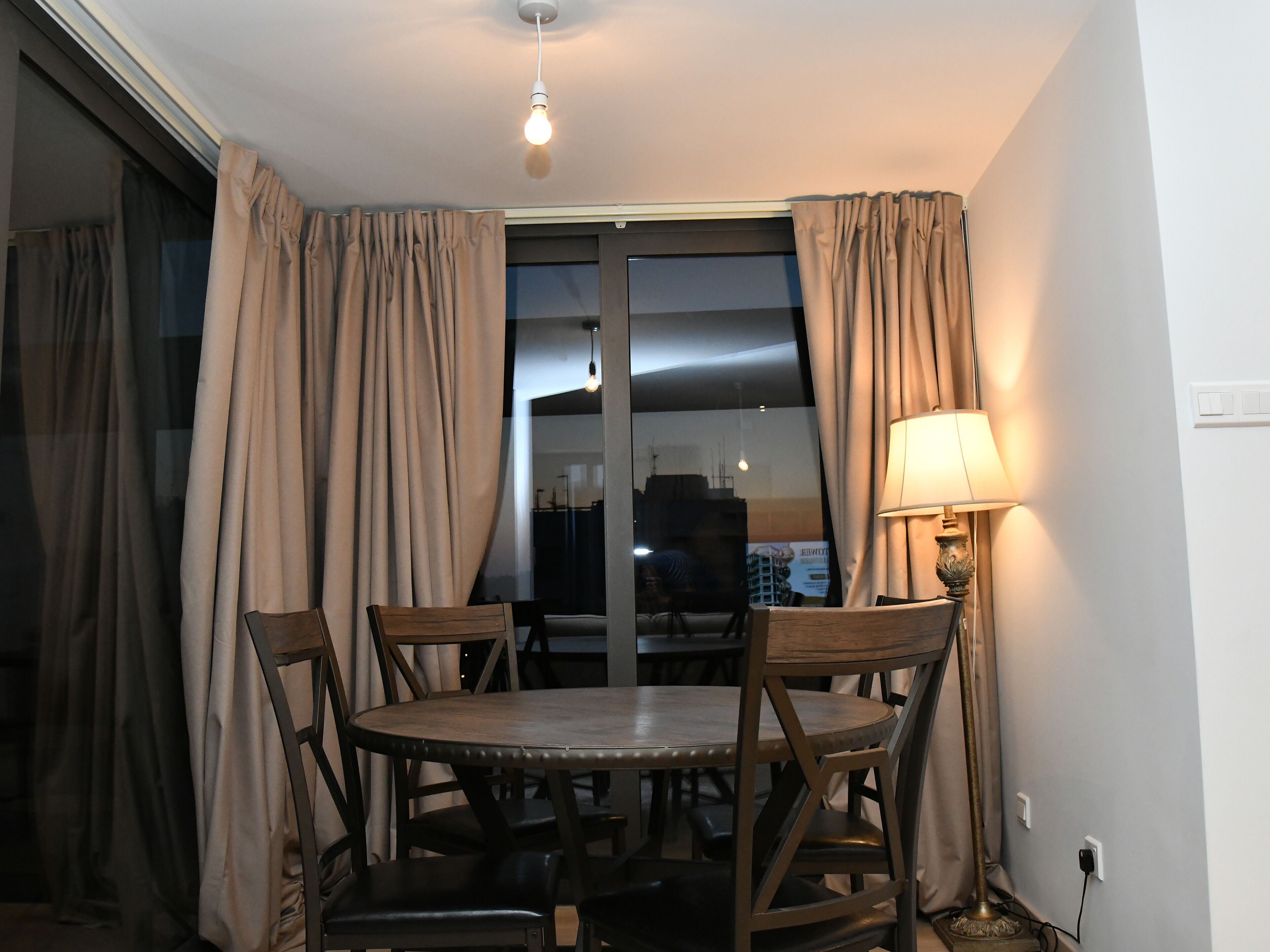 Un comedor con una mesa redonda, cuatro sillas, una lámpara y vistas a través de una ventana con cortinas.