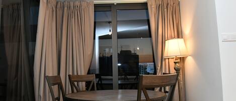 Un comedor con una mesa redonda, cuatro sillas, una lámpara y vistas a través de una ventana con cortinas.