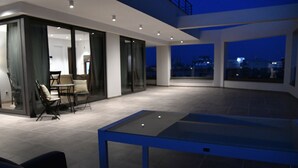Uma área de estar ao ar livre moderna, com mesa e cadeiras, janelas amplas e vista para a paisagem urbana ao anoitecer.