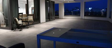 A modern outdoor patio area with a dining table and chairs, large windows, and a view of the cityscape at dusk.