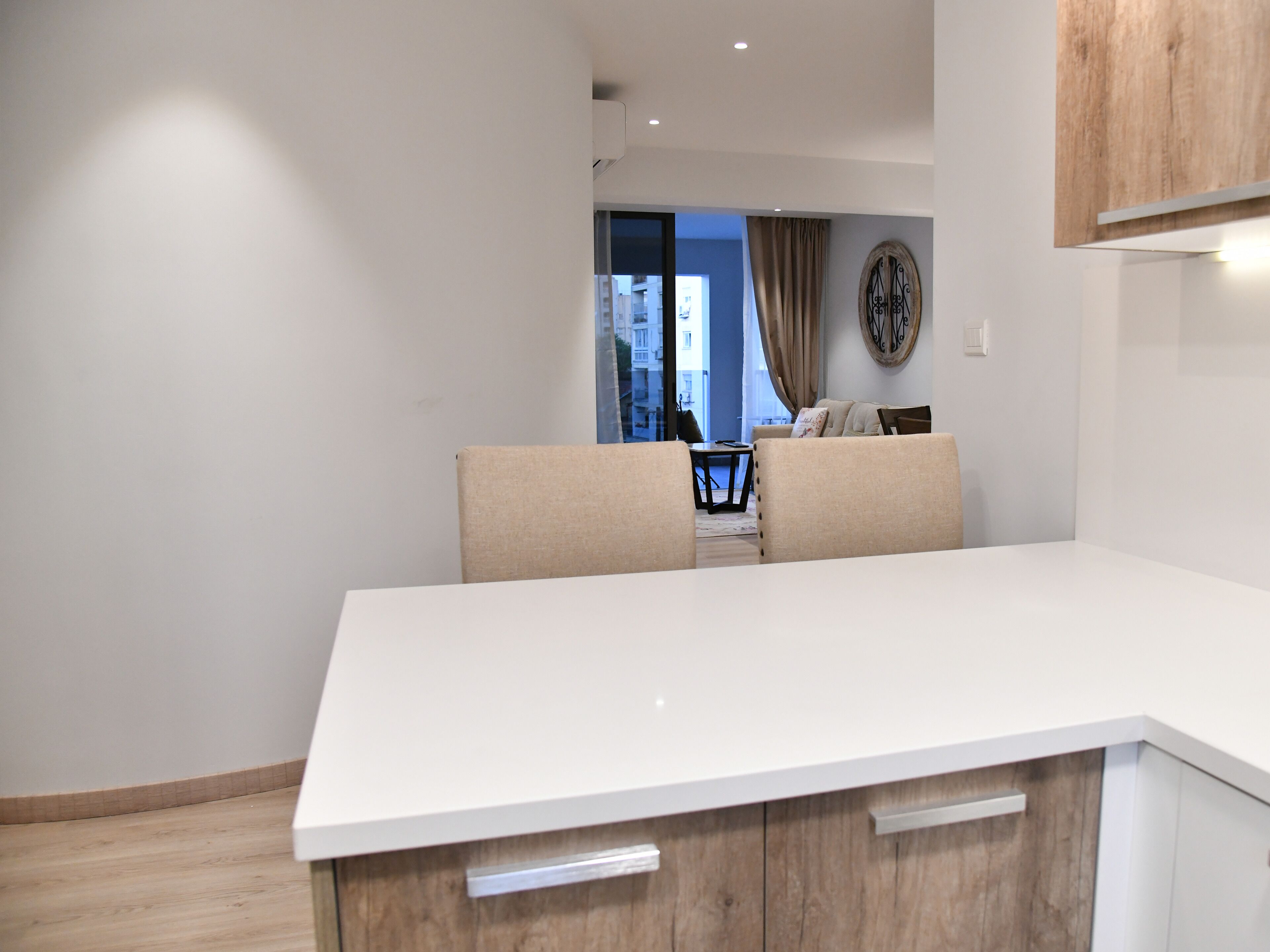 A modern kitchen with white cabinetry and a large countertop. There is a dining area with a table and chairs visible through an open doorway.