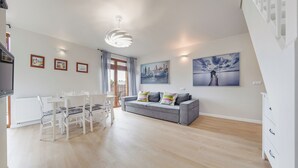 Ein modernes Wohnzimmer mit Essbereich, ein graues Sofa mit bunten Kissen, ein weißer Esstisch mit Stühlen und ein großes Fenster mit Vorhängen.