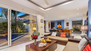 A modern living room with a large sofa, a coffee table, and a dining area. There is a sliding glass door leading to a pool area.