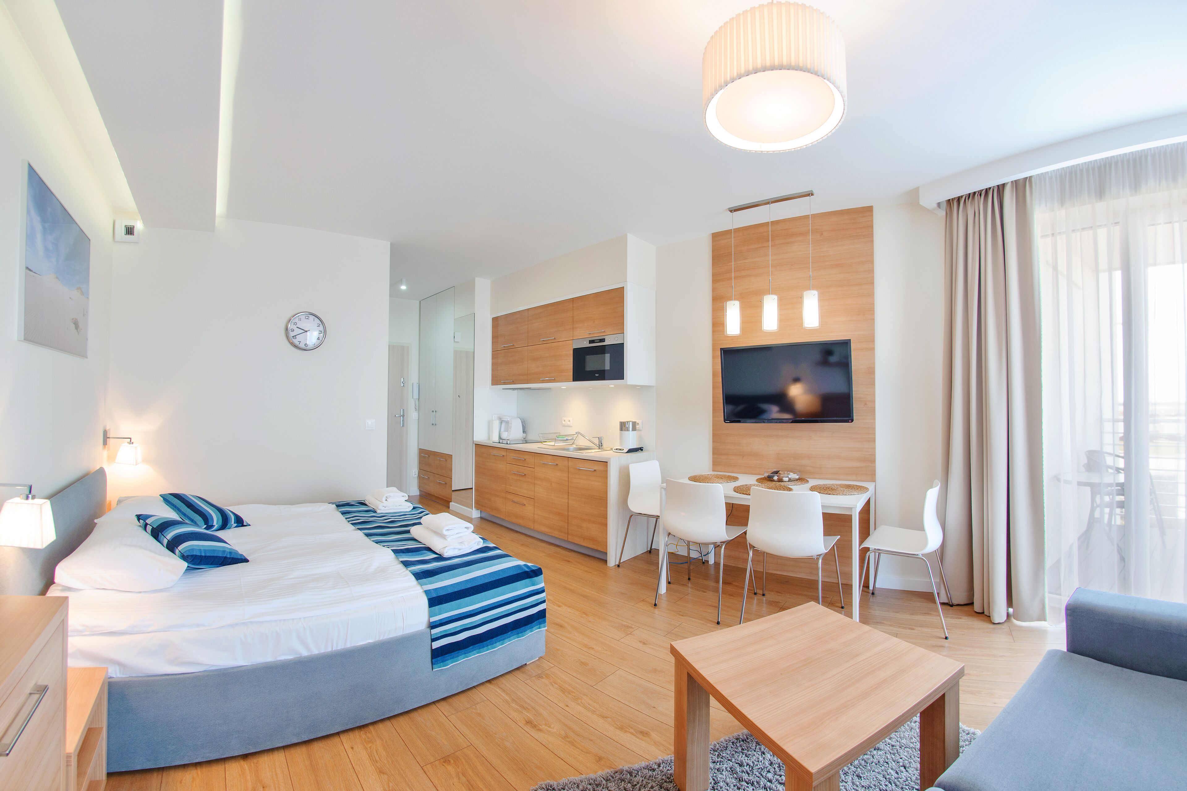 Una habitación de hotel moderna con cama, mesita de noche y un pequeño escritorio con silla. Se aprecia una kitchenette al fondo con microondas y televisor montado en la pared.