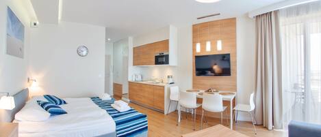 Una habitación de hotel moderna con cama, mesita de noche y un pequeño escritorio con silla. Se aprecia una kitchenette al fondo con microondas y televisor montado en la pared.
