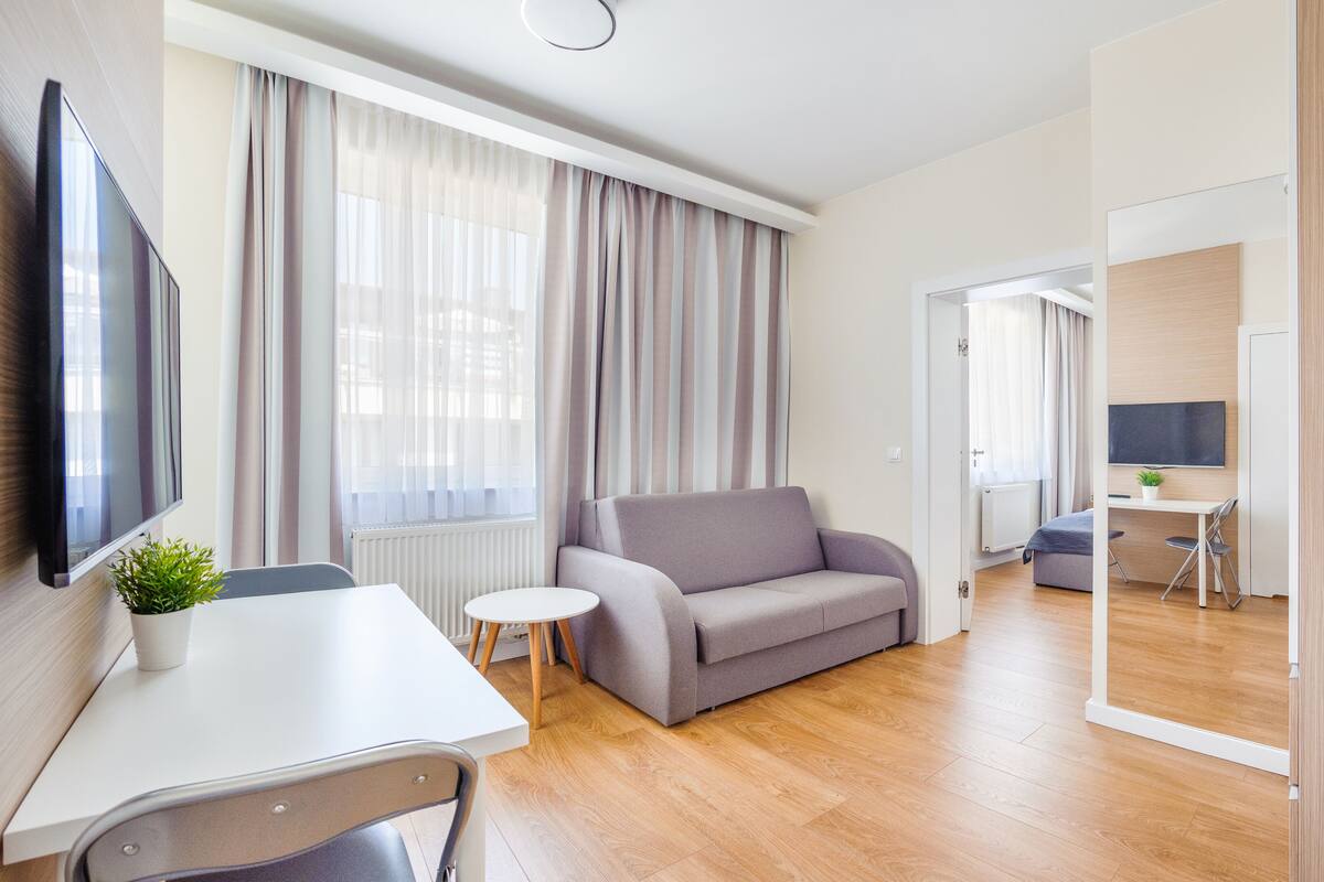 A modern hotel room with a sofa, a small round table, a TV, and a view of the adjacent room.