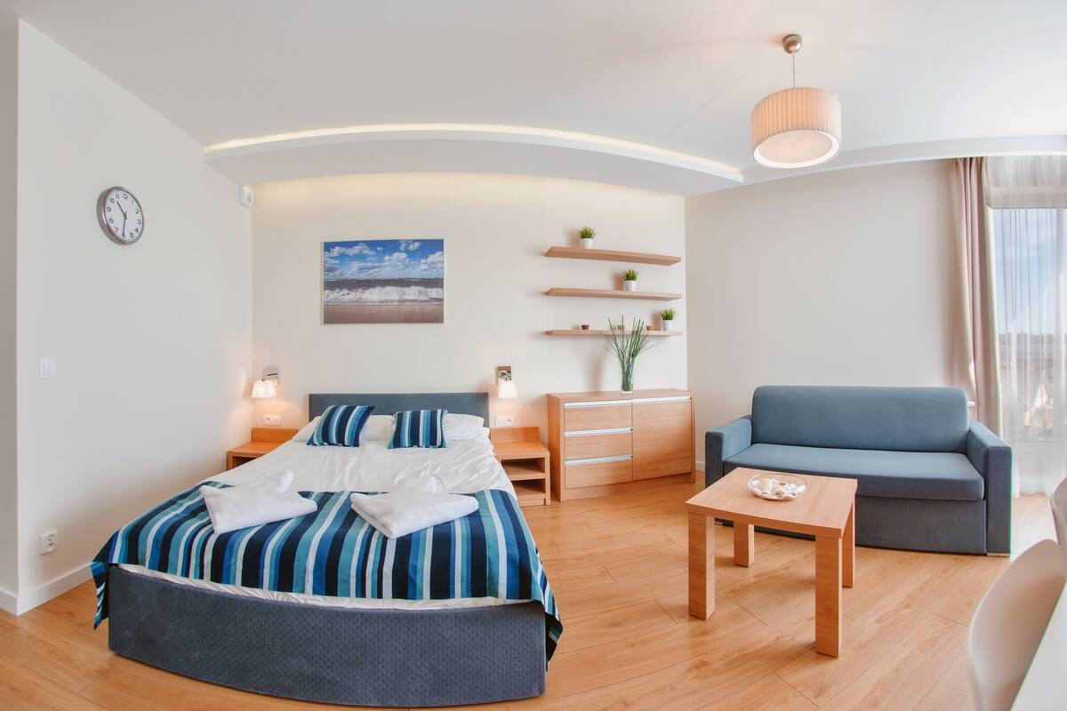 A modern hotel room with a bed, a sofa, a desk, and a small table. There is a clock on the wall and a painting above the bed.