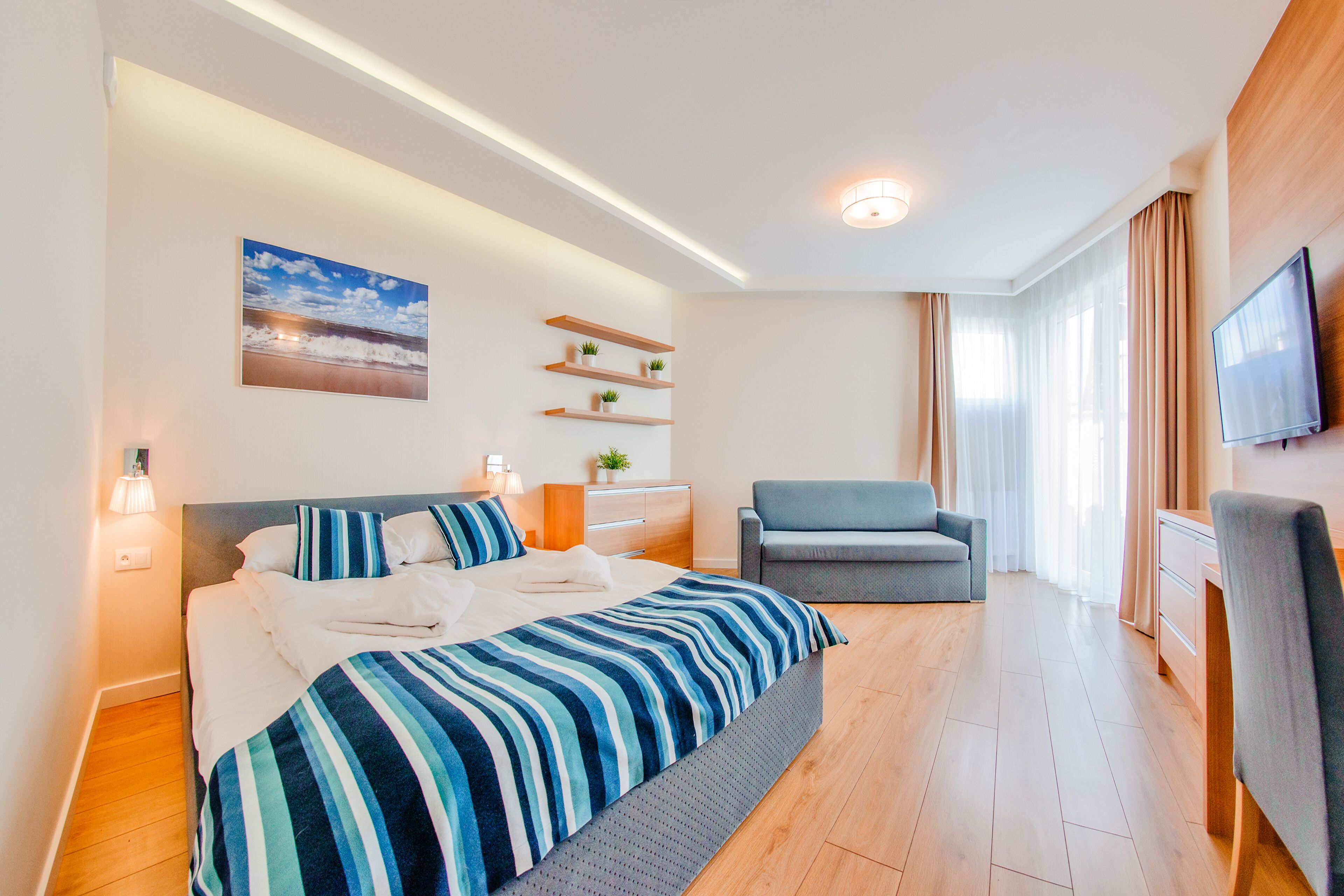 Une chambre d’hôtel moderne avec un grand lit, un canapé gris, un téléviseur fixé au mur et une petite plante posée sur une étagère.