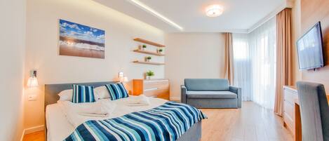 Une chambre d’hôtel moderne avec un grand lit, un canapé gris, un téléviseur fixé au mur et une petite plante posée sur une étagère.