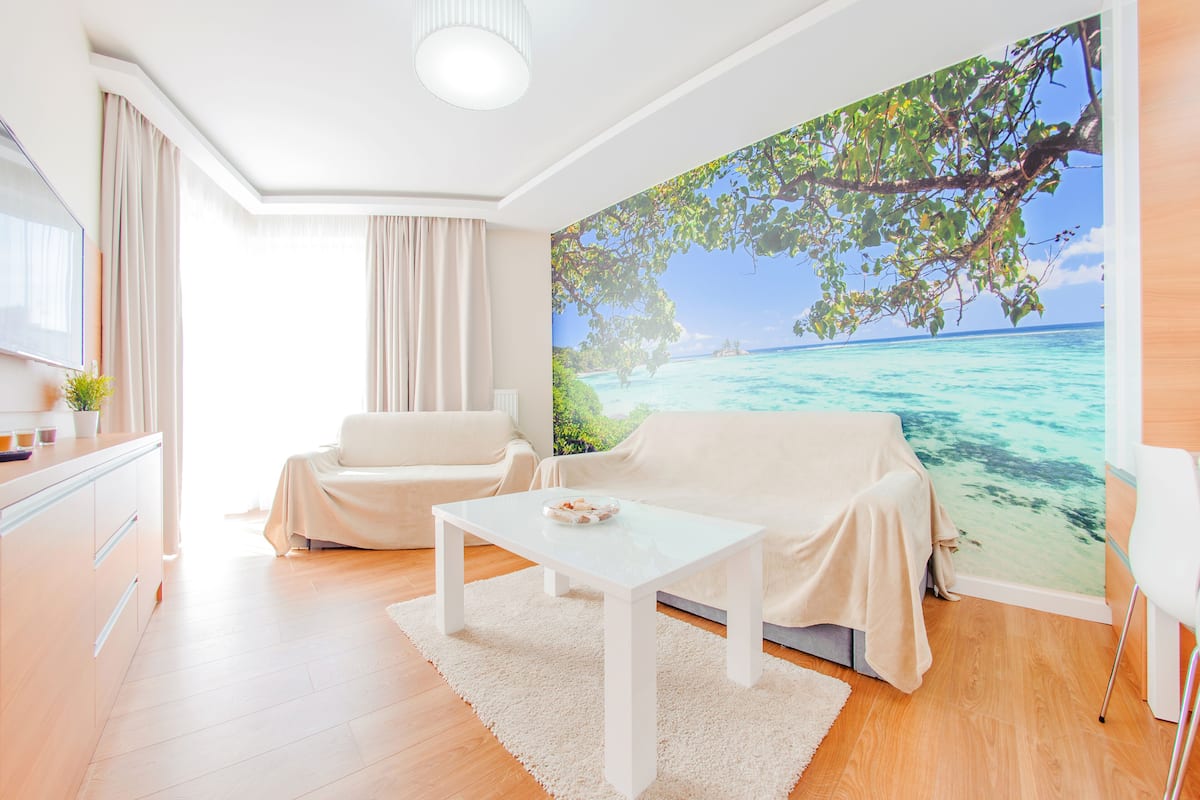 A modern living room with a white sofa, a white coffee table, and a large mural of a tropical beach.