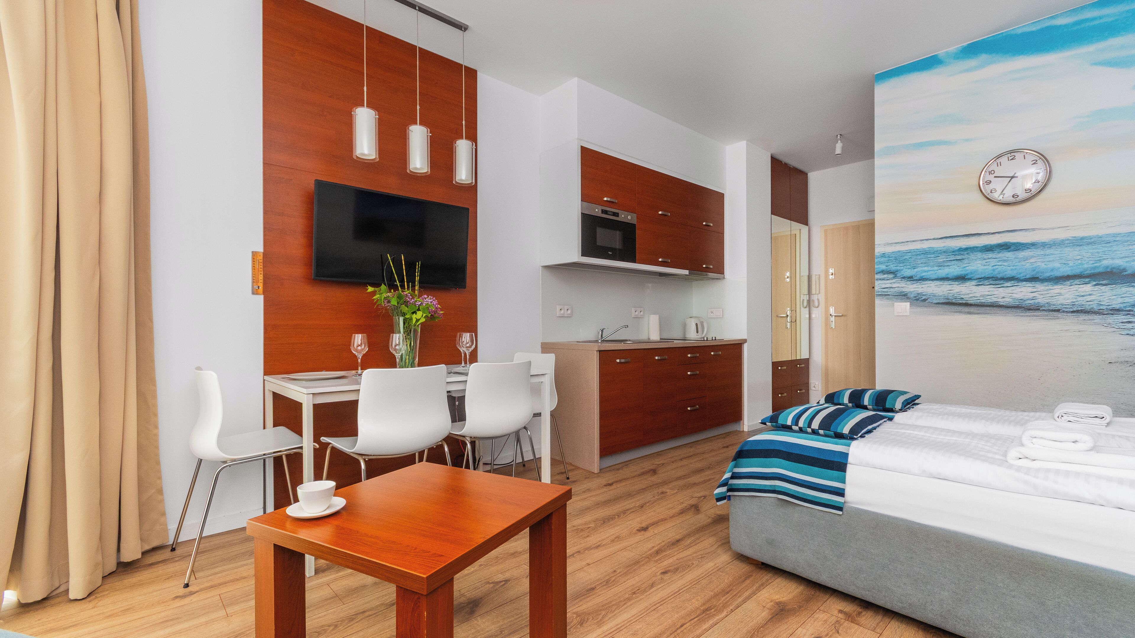 A modern hotel room with a wooden dining table, white chairs, a bed with striped bedding, a TV mounted on the wall, and a kitchen area.