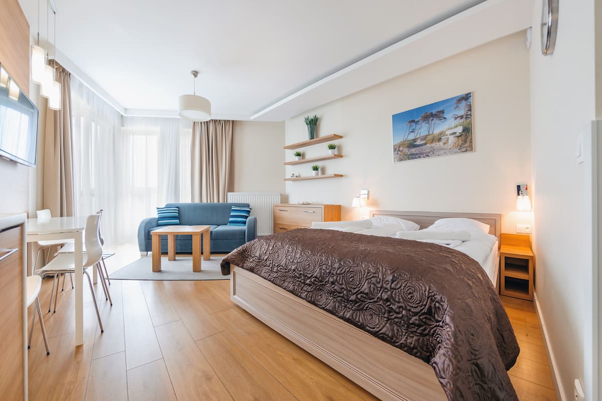 A modern hotel room with a large bed, a desk, a chair, a sofa, and a small table. There are shelves with plants on the wall and a painting above the bed.