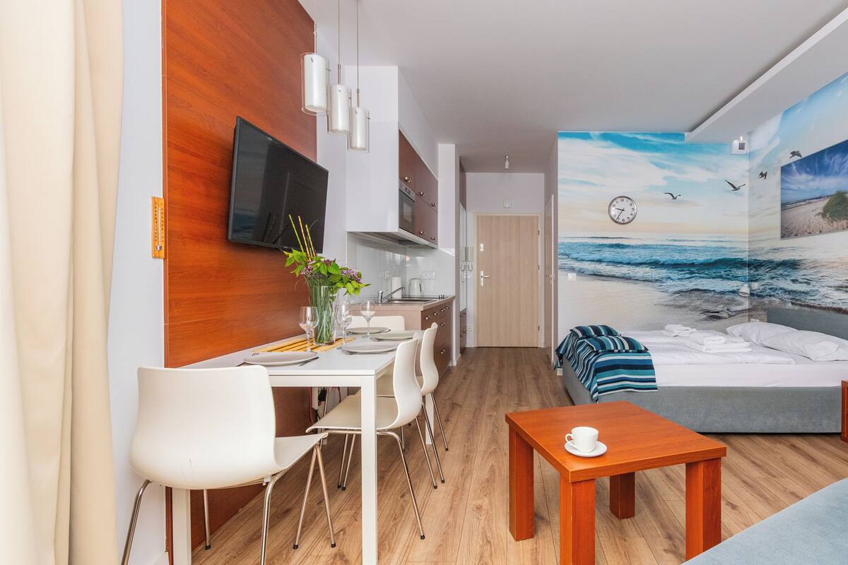 A compact hotel room with a bed, a dining area with a table and chairs, a small kitchen area, and a wall mural of a beach scene.