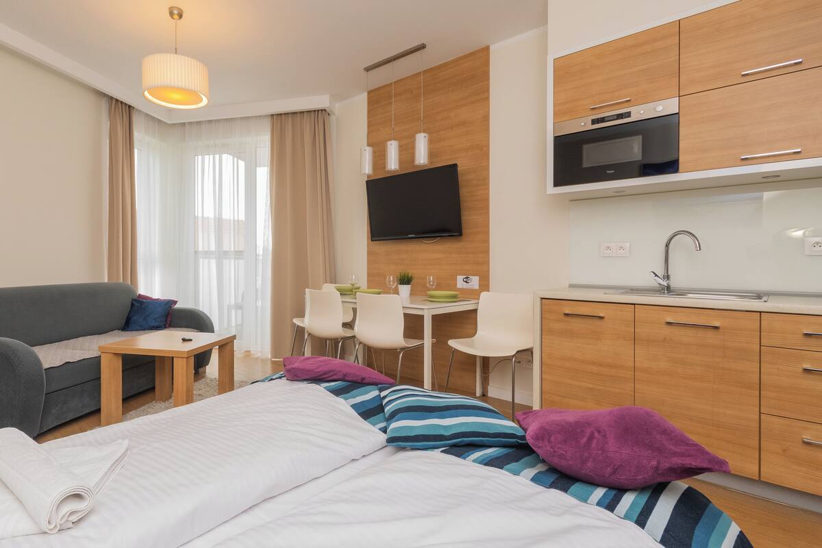 A modern hotel room with a bed, a sofa, a small dining table with chairs, a kitchen area with a microwave and sink, and a TV mounted on the wall.