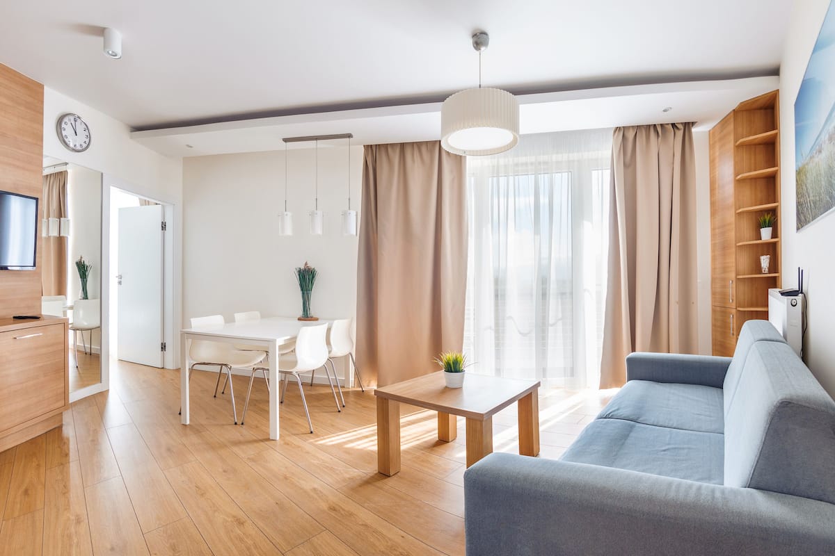 A modern living room with a grey sofa, wooden flooring, a dining area with a white table and chairs, a TV mounted on a wooden cabinet, and a large window with curtains.