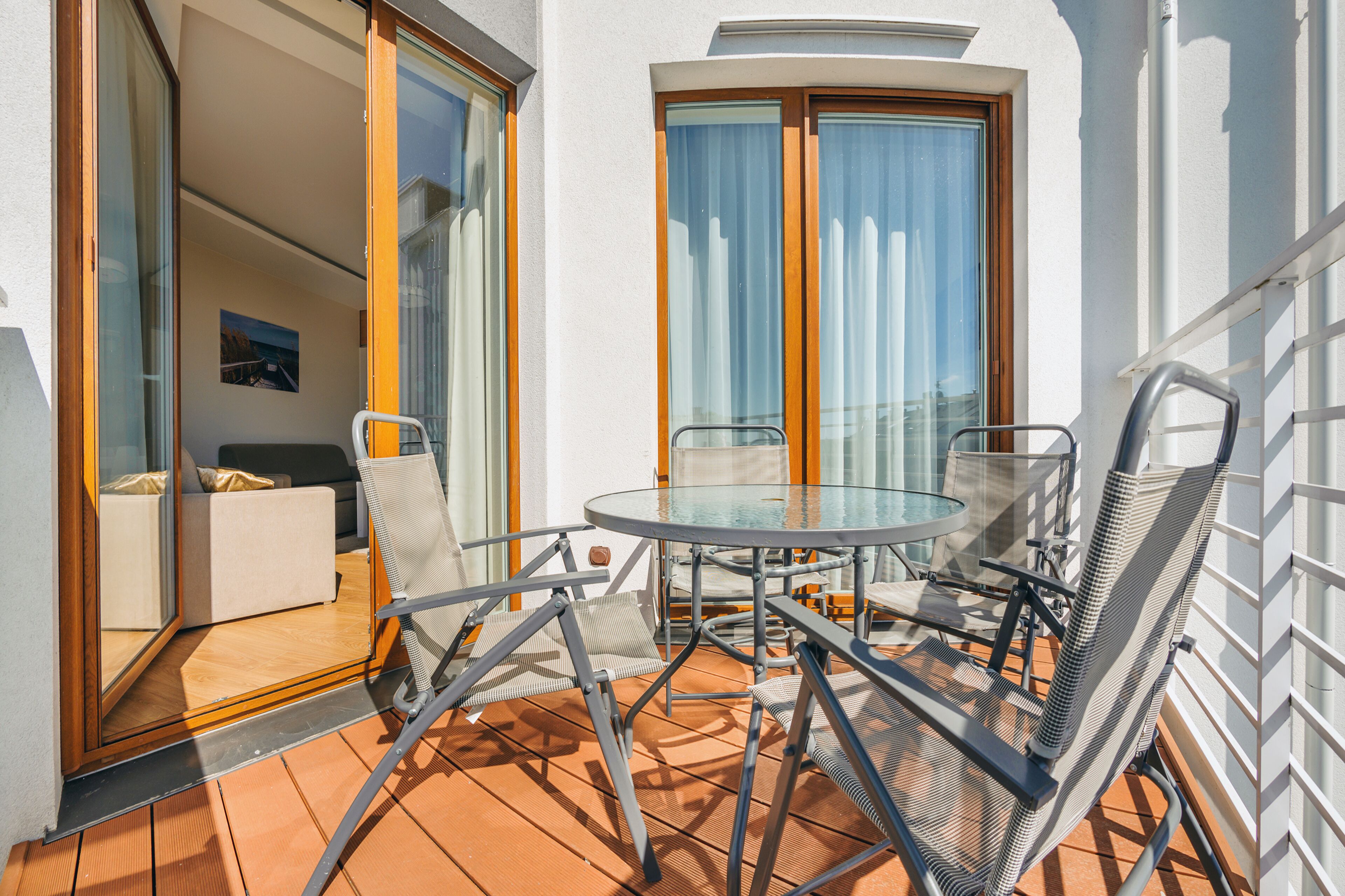 An outdoor balcony with a glass table and metal chairs, a sliding glass door leading to a room, and a view of the sky.