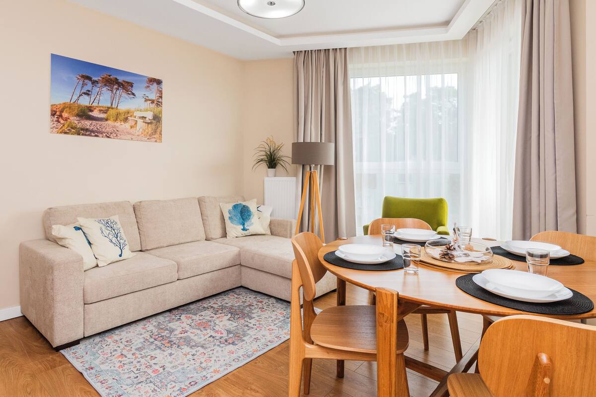 A modern living room with a beige sofa, a dining table set for four, and a large window with sheer curtains.