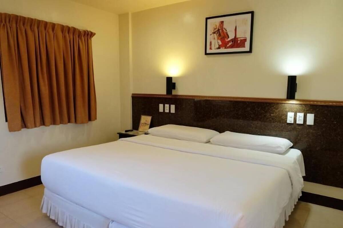 Una habitación de hotel con una cama grande, una mesita de noche, un cuadro enmarcado en la pared y una ventana con cortinas.