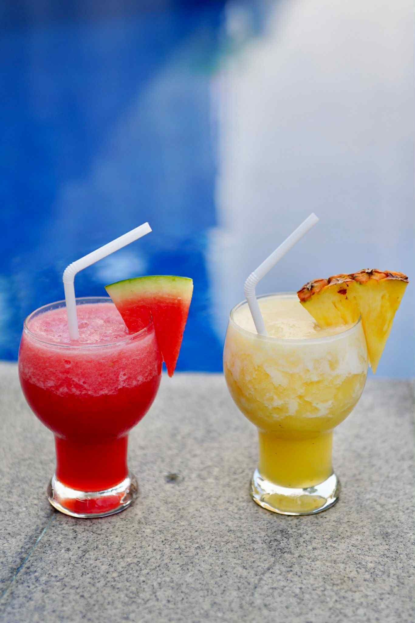 Two drinks with straws and fruit garnishes on a table by a pool.