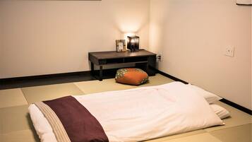 A room with tatami mats, a bed with a white and brown blanket, a small table with a lamp, and a cushion.