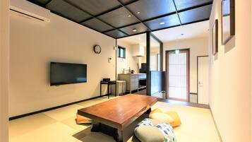 A modern interior with a wooden dining table, tatami mats, a flat-screen TV, and a kitchen area.