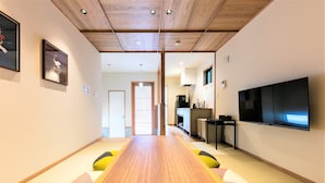 A modern interior with a long wooden table and cushioned seating, a kitchen area, and a flat-screen TV mounted on the wall. - Musubi Hotel Machiya Naraya-machi 2 (Fukuoka)