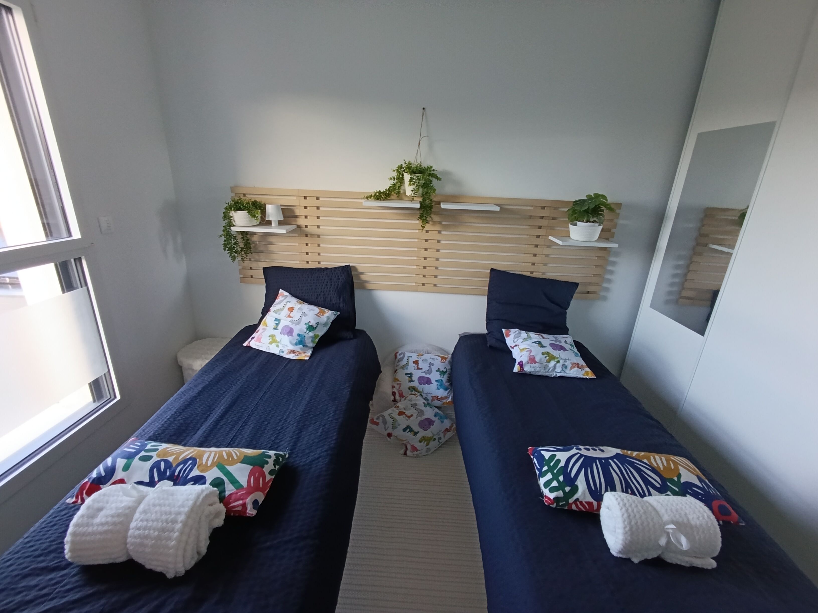 Une petite chambre moderne avec deux lits simples, chacun agrémenté d’oreillers colorés et d’une serviette blanche. Des plantes en pot sont disposées sur une étagère en bois au-dessus des lits, et une fenêtre se trouve sur le côté gauche.