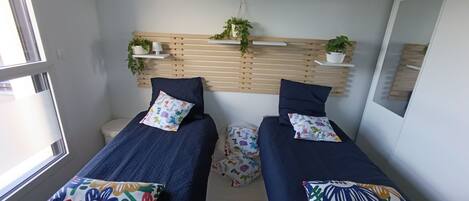 Une petite chambre moderne avec deux lits simples, chacun agrémenté d’oreillers colorés et d’une serviette blanche. Des plantes en pot sont disposées sur une étagère en bois au-dessus des lits, et une fenêtre se trouve sur le côté gauche.