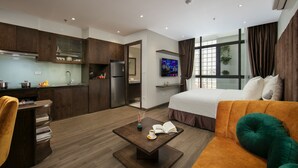 Una habitación de hotel moderna con kitchenette, una cama con ropa de cama verde y blanca, una mesita de centro con un libro y un florero, y un ventanal con cortinas.
