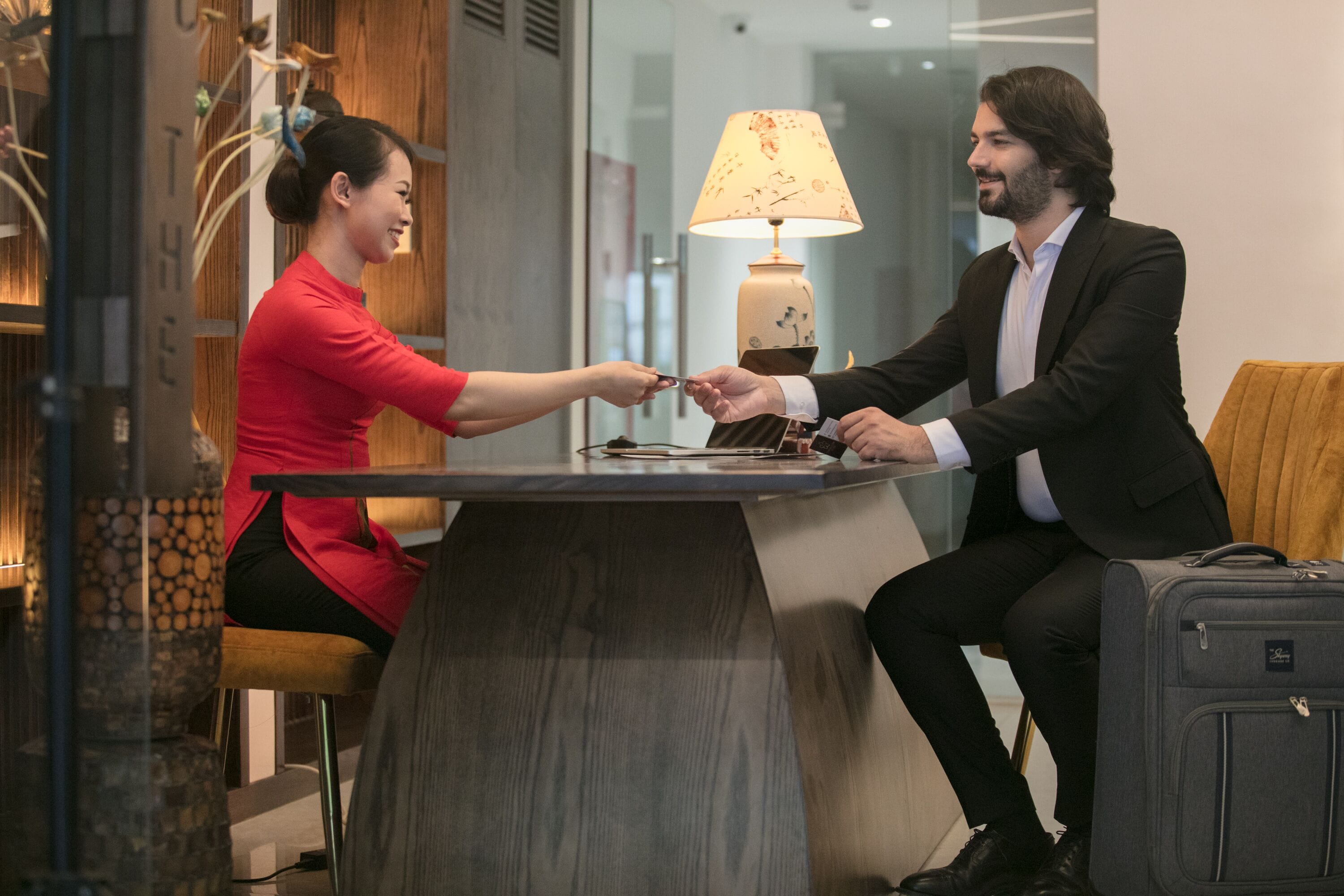 A man and a woman seated at a table with a lamp, in a room with a wooden accent wall and a suitcase on the floor.