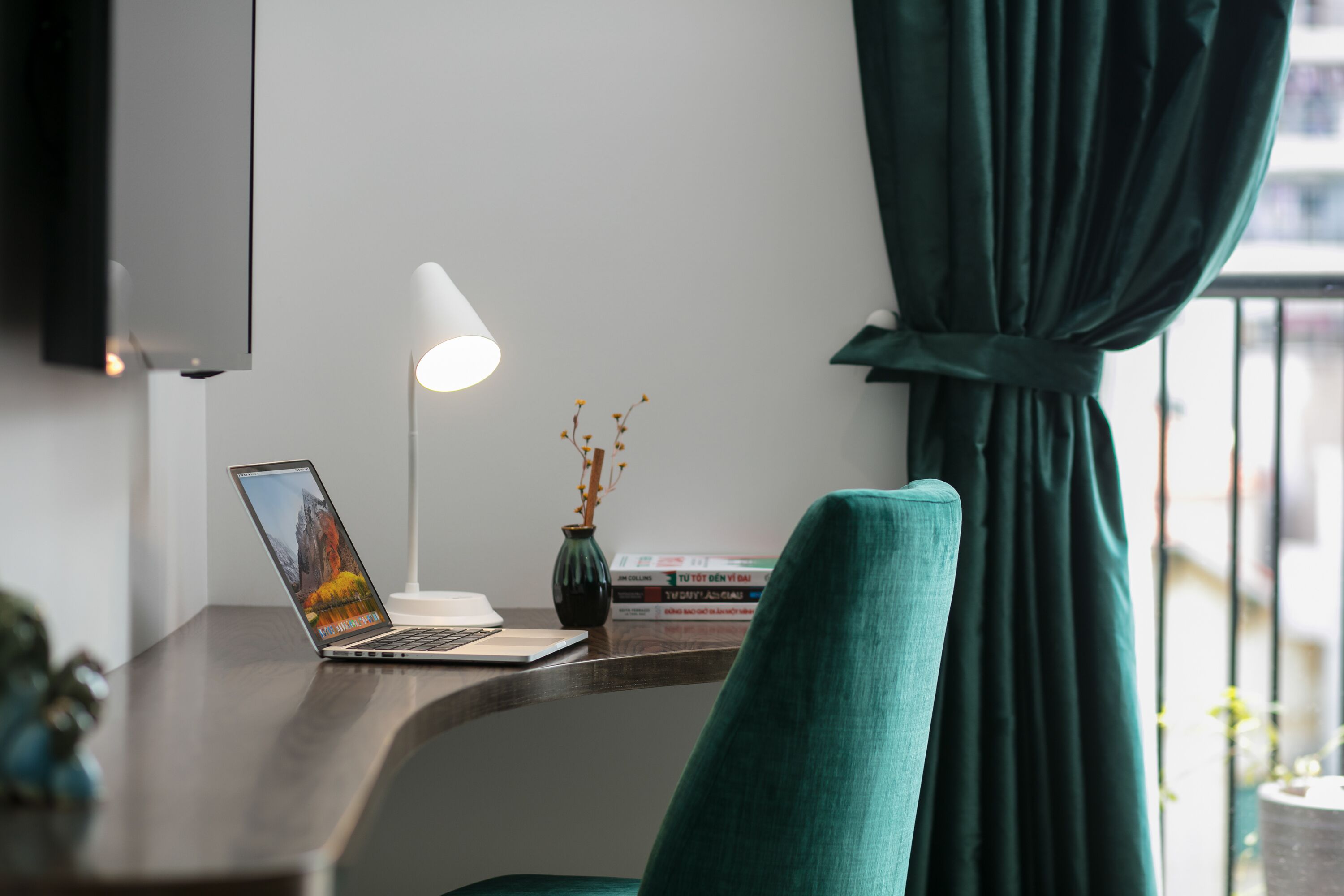 A modern workspace with a laptop, a white desk lamp, a vase with dried branches, and a teal chair.