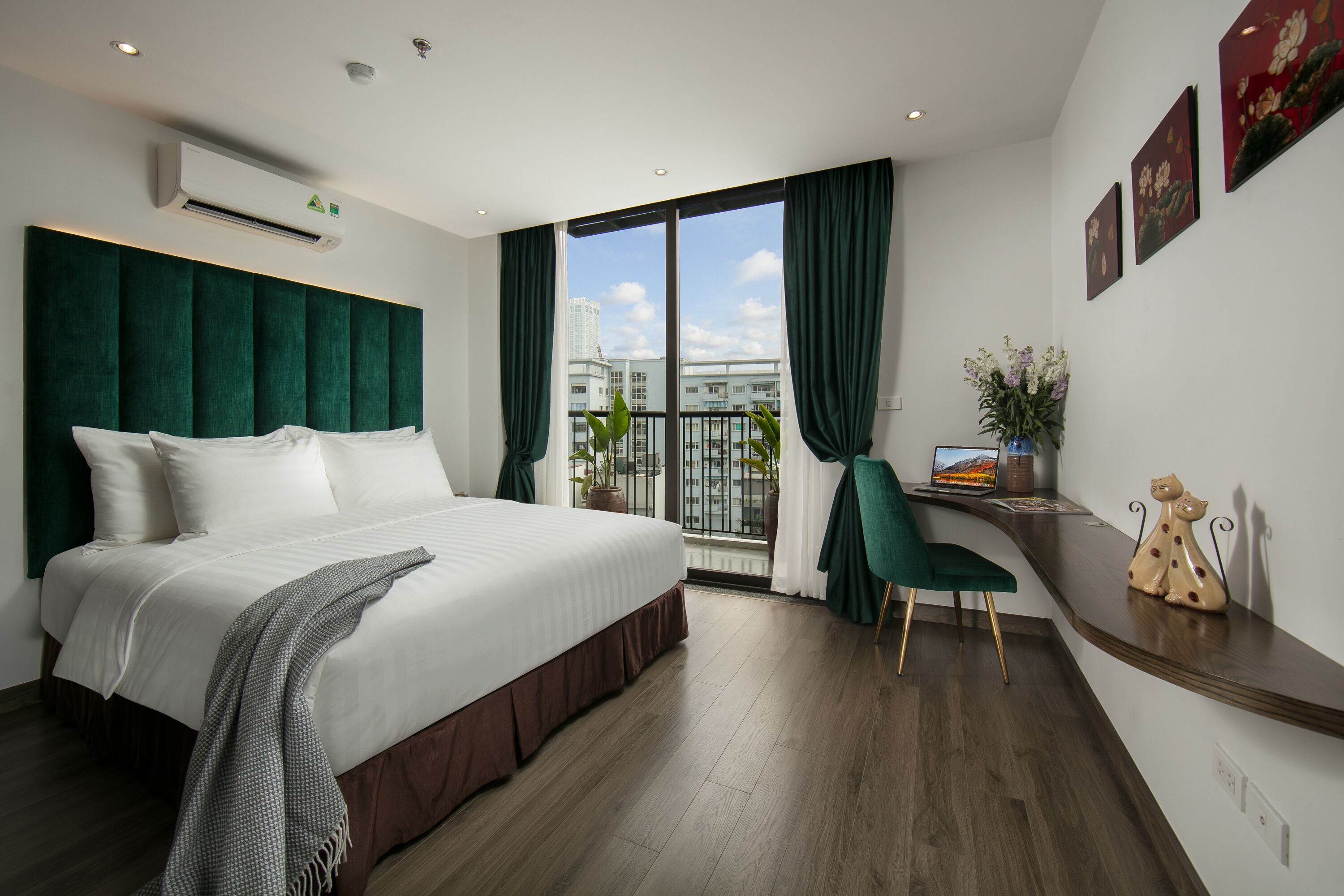 Habitación de hotel moderna con una cama grande, un escritorio con una computadora portátil, vistas a la ciudad y un balcón con plantas.