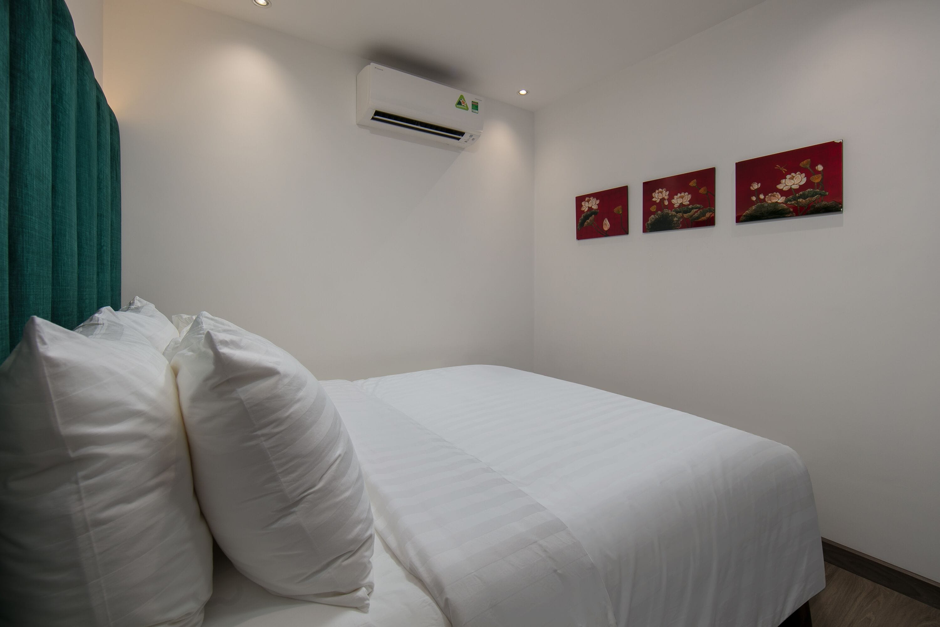 A neatly made bed with white bedding and pillows, a wall-mounted air conditioner, and three red floral paintings on the wall.