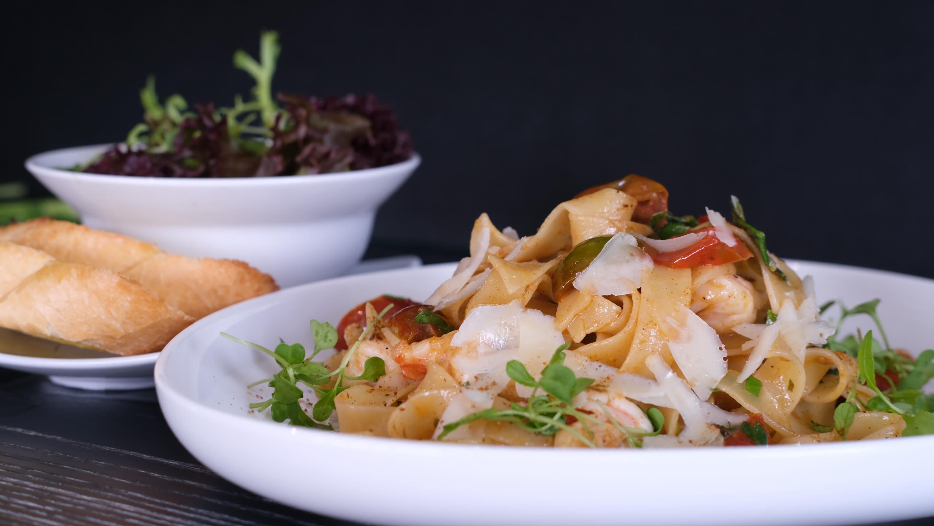 Un plato de pasta con vegetales y queso, un bol de ensalada y un trozo de pan sobre una mesa.