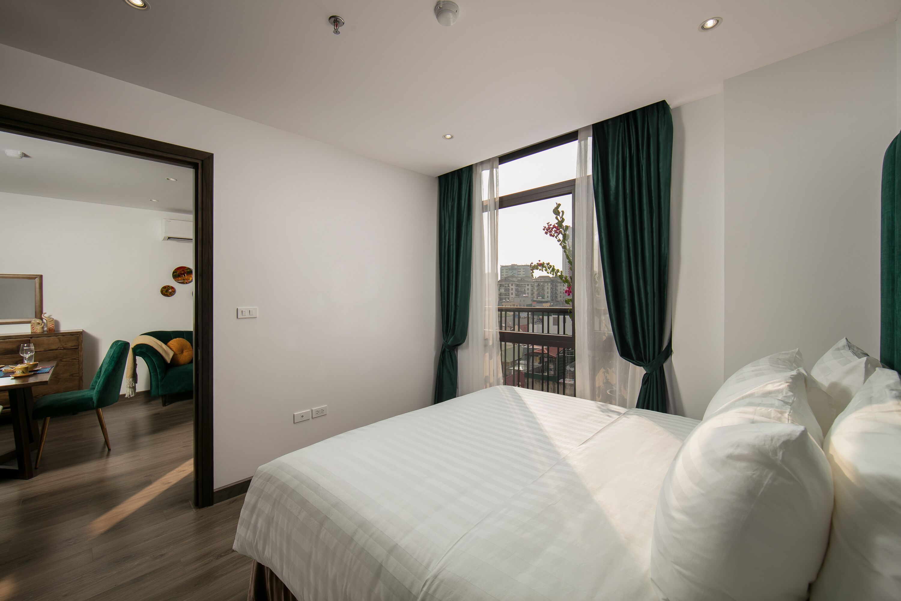 Una habitación de hotel moderna con una cama grande, un escritorio con una silla y vistas a edificios a través de una ventana con cortinas verdes.