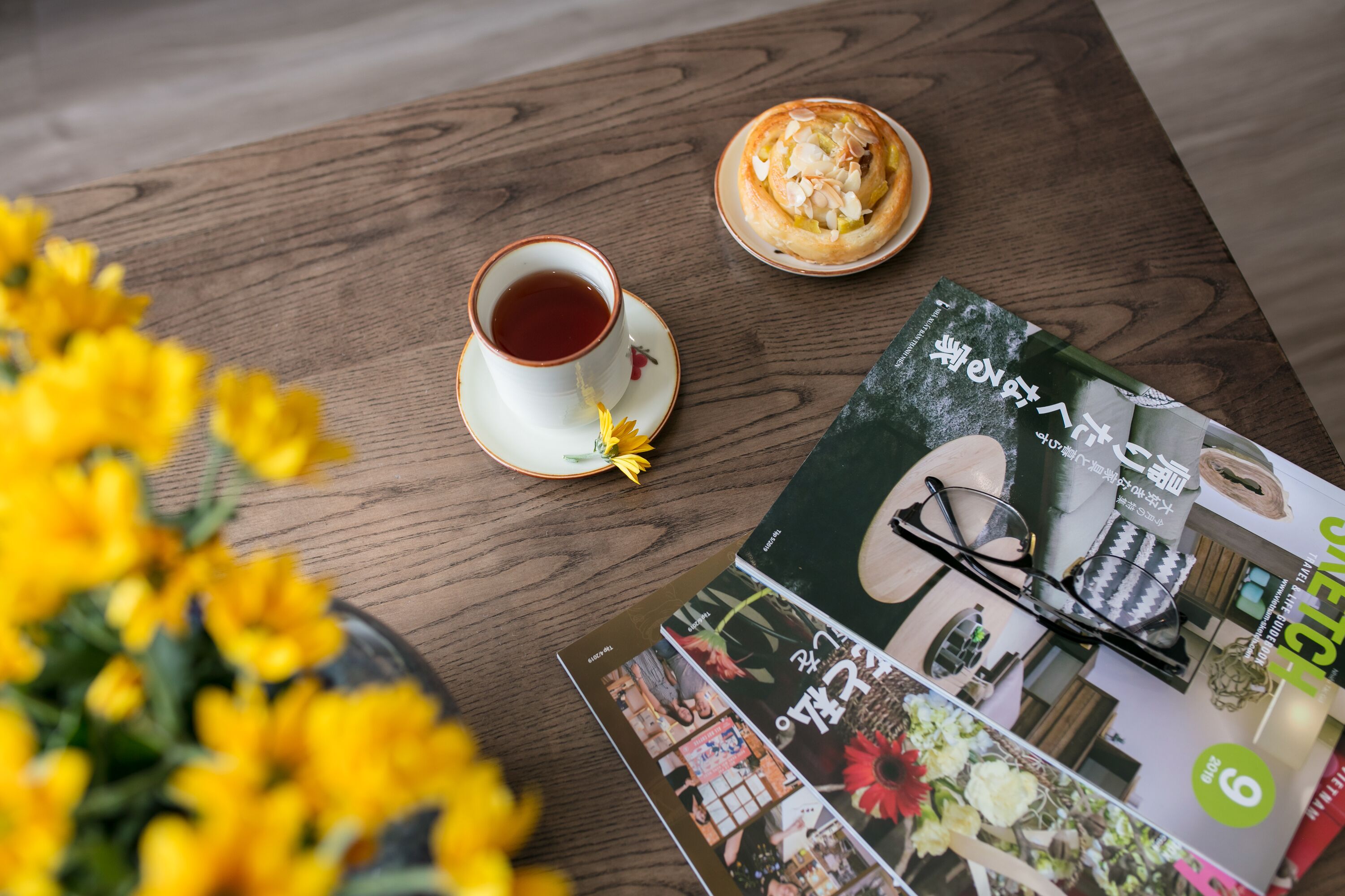 Una mesa de madera con una taza de té, un pastel, una revista y un par de gafas.
